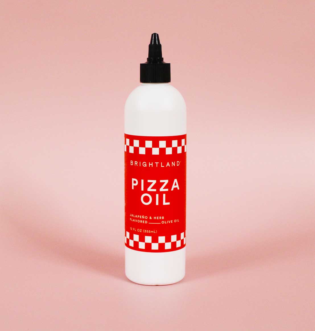 Brightland Pizza Oil bottle standing against a soft pink background. The bold red label with a checkered design highlights the jalapeño and herb-infused olive oil, crafted for drizzling on pizza, pasta, and more. Modern and minimal product shot emphasizing premium quality.