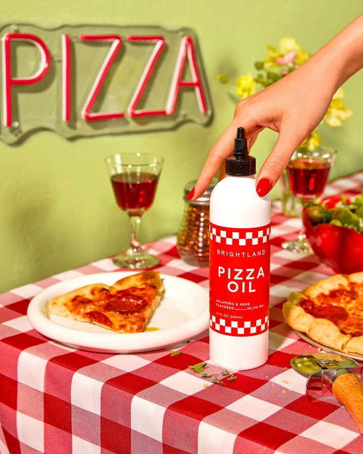 Brightland Pizza Oil bottle on a red and white checkered tablecloth, surrounded by fresh pepperoni pizza, wine glasses, and a retro ‘PIZZA’ neon sign. A hand with red nail polish holds the bottle, highlighting the spicy jalapeño and herb-infused olive oil. Perfect for drizzling on pizza, pasta, and more.