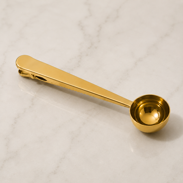 A polished gold-colored metal measuring spoon with a long handle and a spring-loaded clip at the end, resting on a white marble surface.