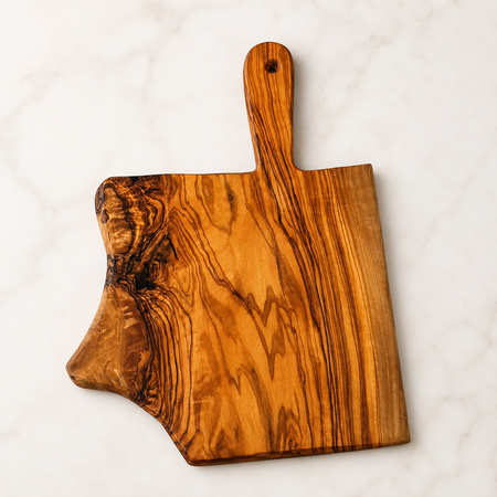 An olive wood serving board with a handle and a natural irregular shape, displayed on a white marble surface.